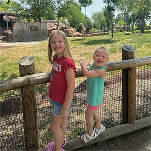 Two little girls standing by fence with animals in the background